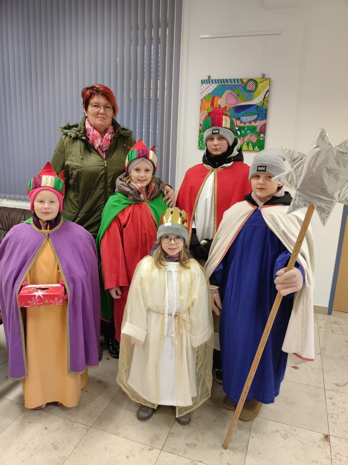 Eine Gruppe von Kindern und einer erwachsenen Frau posiert für ein Foto in einem Raum. Sie sind alle in Kostümen gekleidet, möglicherweise für ein Theaterstück. Sie stehen in einer Reihe, wobei eine Frau einen Sternenstab hält. Hinter ihnen befindet sich eine Wand mit einem Gemälde.