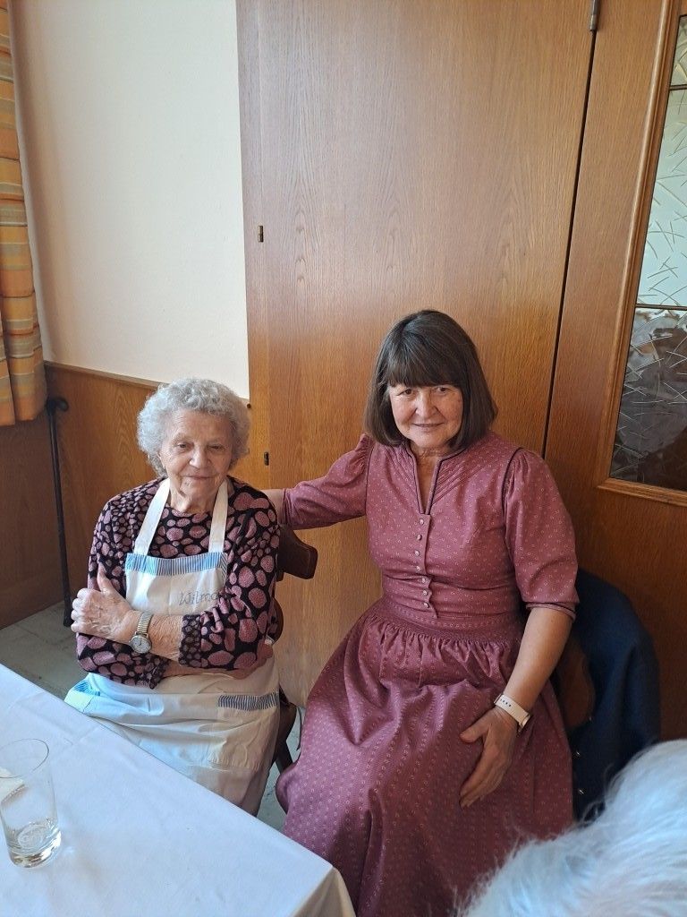 Zwei Frauen sitzen in Stühlen an einem Tisch, eine in einem rosa Kleid und die andere in einer gepunkteten Schürze. Sie lächeln und posieren für ein Foto.