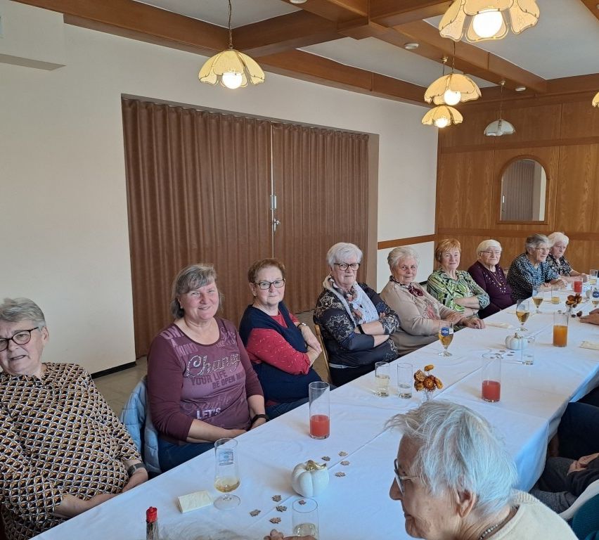 Eine Gruppe älterer Frauen sitzt an einem langen Tisch mit Getränken und Dekorationen in einem Raum mit Holzwänden und Deckenlampen.