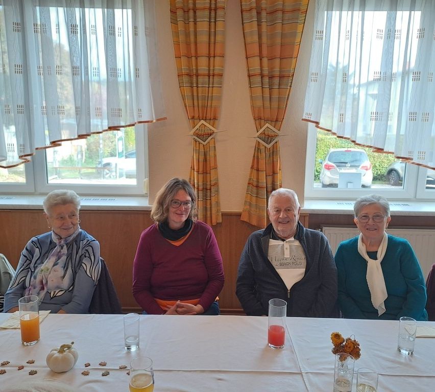 Vier ältere Erwachsene sitzen lächelnd am Tisch mit Getränken und Kürbissen. Hinter ihnen haben zwei Fenster Vorhänge. Draußen sind Autos vor Bäumen geparkt.