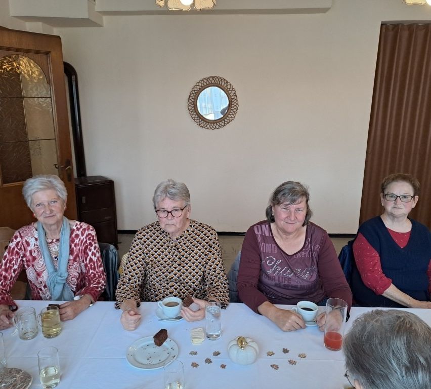 Fünf ältere Frauen sitzen an einem Tisch mit Tassen, Tellern und herbstlichen Dekorationen. Der Raum scheint ein Esszimmer mit weißer Wand, einem runden Spiegel und einer Holztür zu sein.