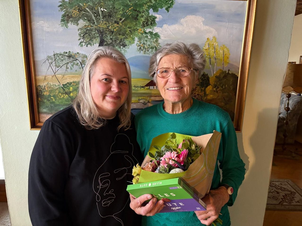 Zwei Frauen posieren vor einem Landschaftsgemälde. Die Frau auf der linken Seite lächelt und trägt ein schwarzes T-Shirt. Die Frau auf der rechten Seite hält einen Blumenstrauß und eine Schachtel 'Mute' Schokoladen.