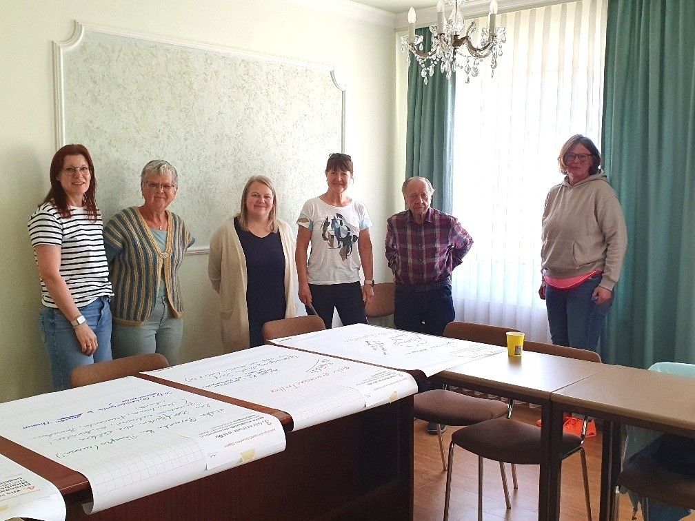 Fünf Erwachsene, zwei Frauen und drei Männer, stehen in einem Raum mit einer weißen Wand, in lässiger Kleidung. Ein Schreibtisch mit Papieren steht vor ihnen. Eine Kronleuchter und eine Lampe hängen von der Decke.
