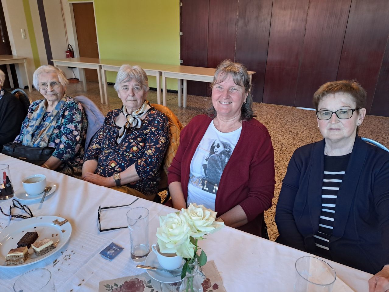 Vier Frauen sitzen an einem Tisch mit weißer Tischdecke und lächeln. Zwei tragen eine Brille, eine hat eine Krawatte und eine andere trägt einen roten Pullover. Es gibt ein Blumenarrangement und Teller mit Essen. Der Hintergrund besteht aus einer gelben Wand und einer braunen Tür.