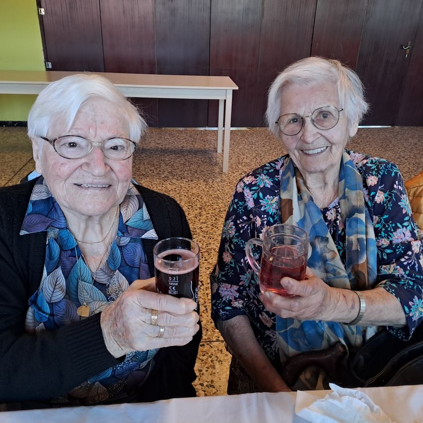 Zwei ältere Frauen sitzen nebeneinander an einem Tisch, lächelnd, mit Gläsern mit roter Flüssigkeit in der Hand. Eine Frau trägt eine Brille und ein blaues Blumenhemd. Die andere trägt einen Schal, eine Armbanduhr und einen goldenen Ring. Hinter ihnen steht eine Holzwand.