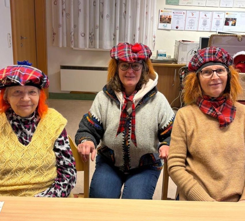 Drei Frauen in Tartanschalen und -tüchern sitzen an einem Tisch und lächeln für ein Foto. Dahinter befindet sich eine Wand mit Papieren und einem Heizkörper.
