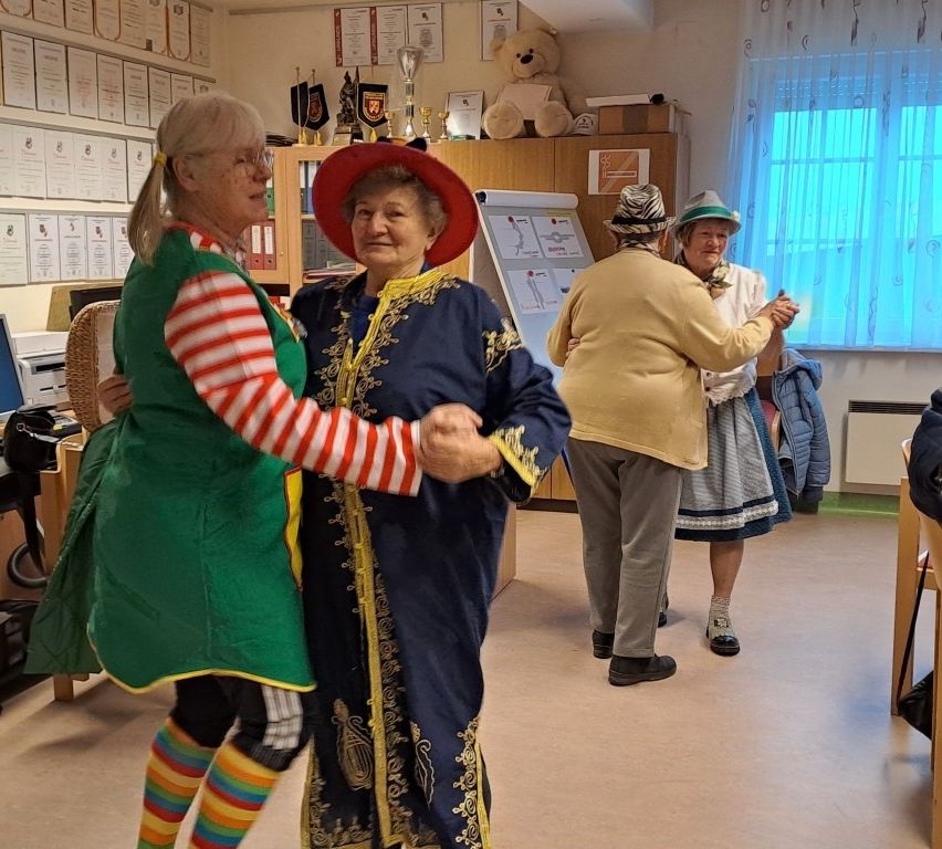 Drei Frauen in einem Raum, zwei tanzen, eine hält ein Brett. Ein Teddybär und Trophäen sind auf einem Regal im Hintergrund.