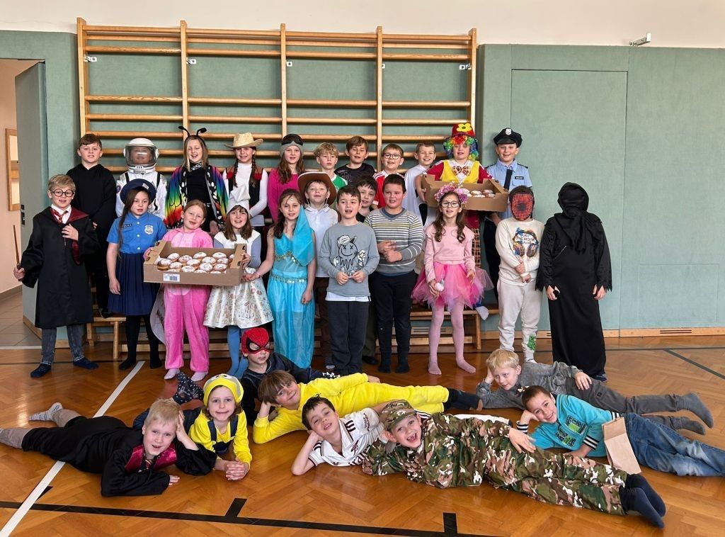 Eine Gruppe von Kindern in verschiedenen Kostümen posiert für ein Foto in einer Turnhalle. Einige halten Requisiten und andere liegen auf dem Boden. Im Hintergrund befinden sich Holzregale.