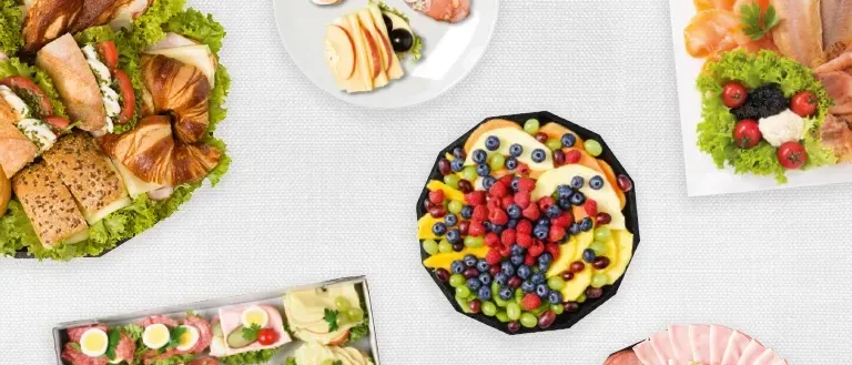 Eine Auswahl an Speisen auf einem Tisch, darunter Sandwiches, Früchte, Fleisch und Salate. Jedes Gericht ist auf einem separaten Teller oder einer Platte.