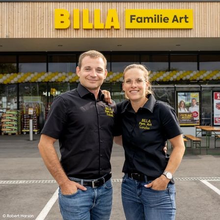 Zwei Personen stehen vor einem BILLA-Geschäft und tragen identische Hemden. Das Geschäft ist mit gelben und weißen Luftballons dekoriert. Hinter ihnen stehen Tische und Bänke, und das Geschäft hat große Fenster.