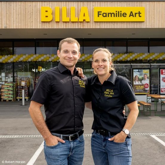 Zwei Personen stehen vor einem BILLA-Geschäft und tragen identische Hemden. Das Geschäft ist mit gelben und weißen Luftballons dekoriert. Hinter ihnen stehen Tische und Bänke, und das Geschäft hat große Fenster.