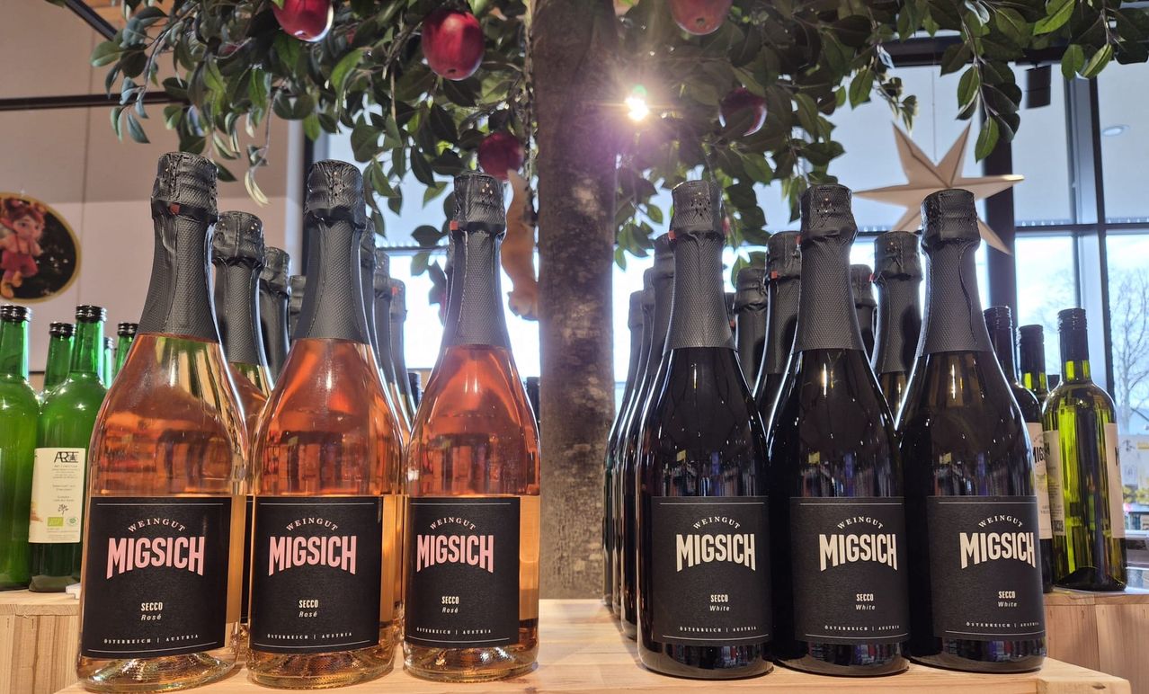 Mehrere Flaschen des Migsich-Weins sind auf einer Holzoberfläche ausgestellt. Die Flaschen haben verschiedene Größen und schwarze Etiketten mit weißem Text. Im Hintergrund ist ein Apfelbaum mit roten Äpfeln zu sehen.