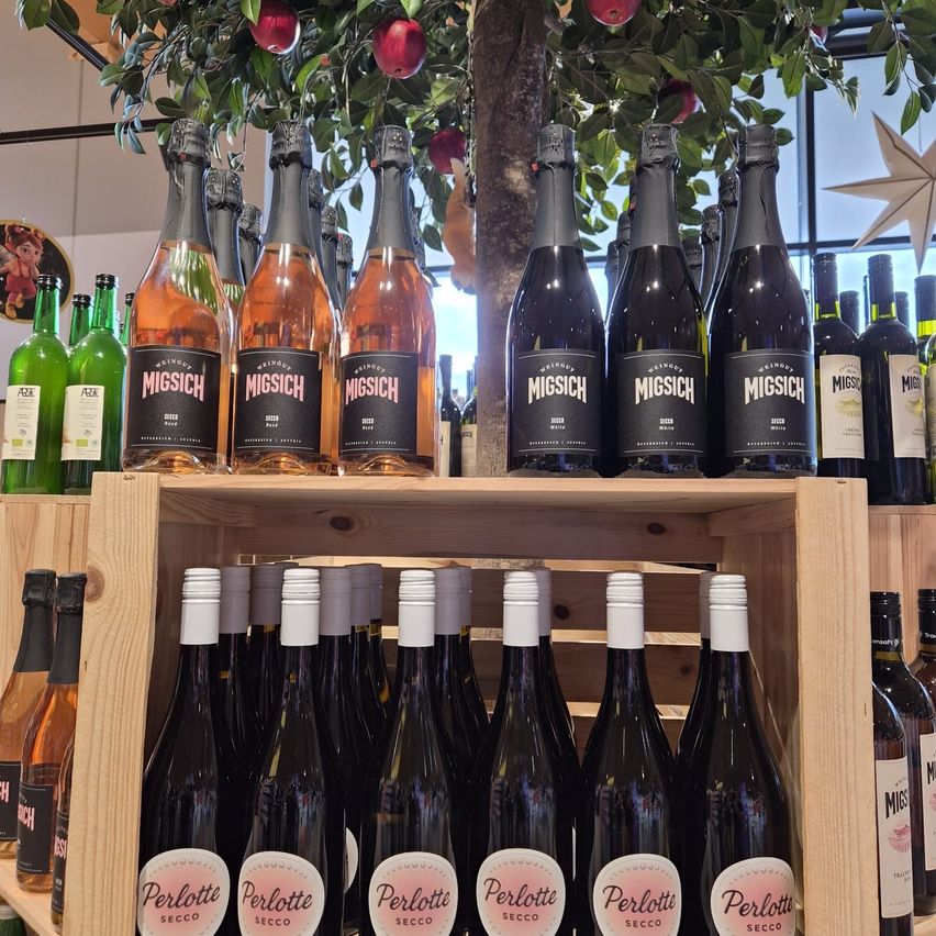 Ein Holzregal zeigt mehrere Flaschen Perlotte Secco und Migisch Wein, mit Äpfeln, die von einem Baum hängen.