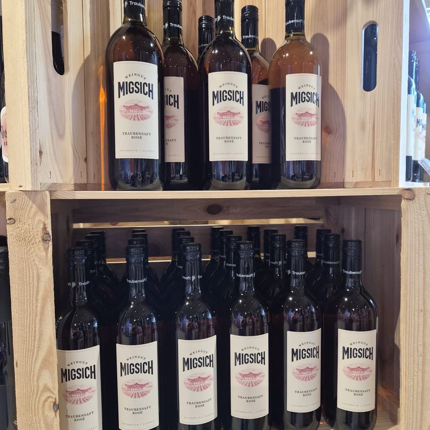 Mehrere Flaschen Wein mit dem Label Migsch sind auf hölzernen Regalen angeordnet. Die Flaschen sind dunkelbraun und einige haben Etiketten mit dem Wort Traubensaft Rose.