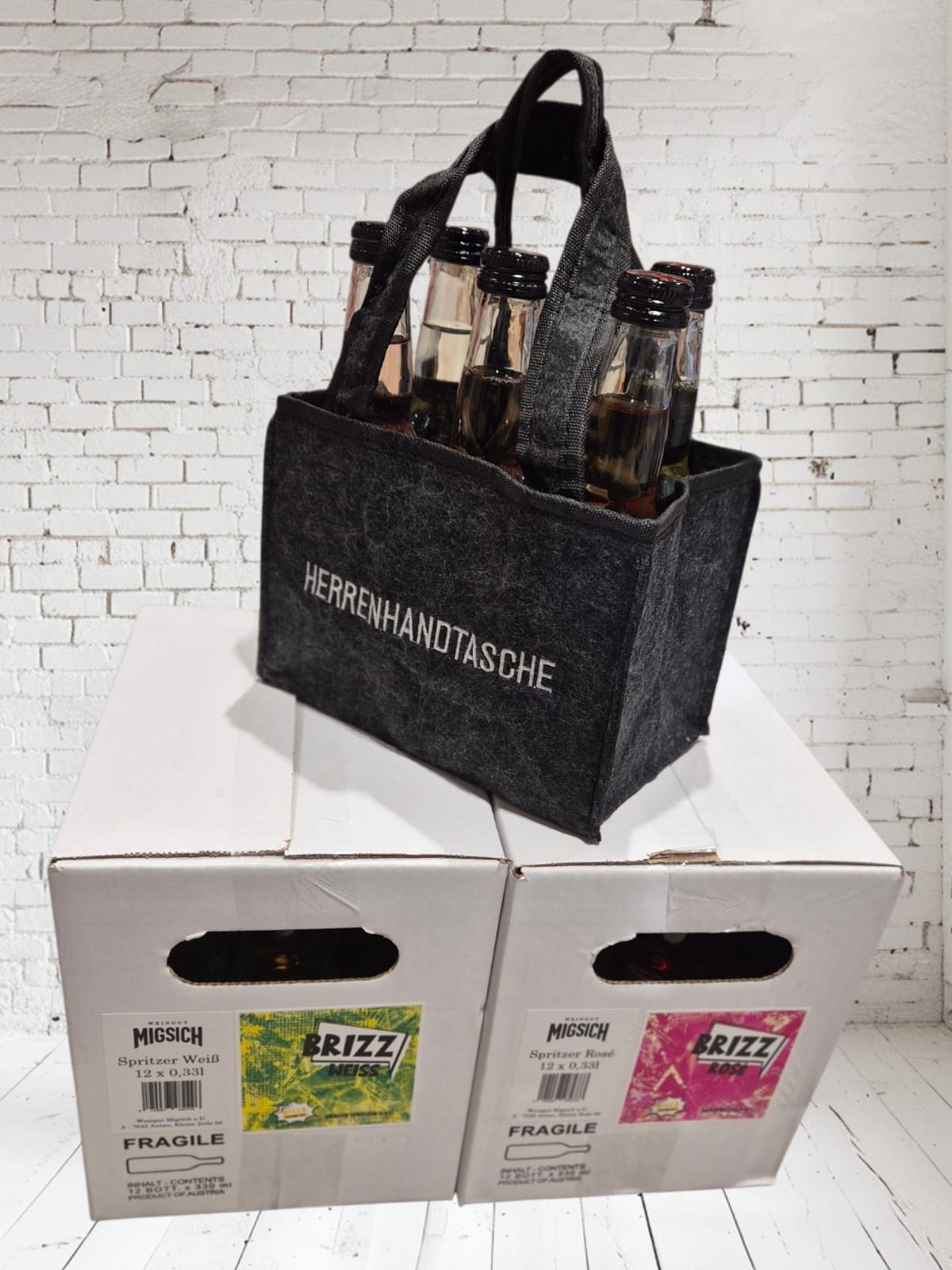 Eine schwarze Filztasche mit dem Aufdruck Herrenhandtasche enthält sechs Bierflaschen. Die Tasche liegt auf zwei weißen Kisten mit der Aufschrift Migisch und Brizz.