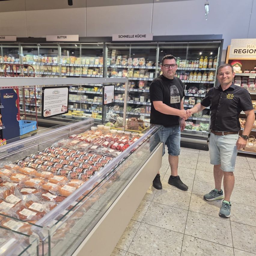Zwei Männer schütteln sich die Hände in einem Supermarkt vor einer Glasvitrine voller Fleischprodukte.