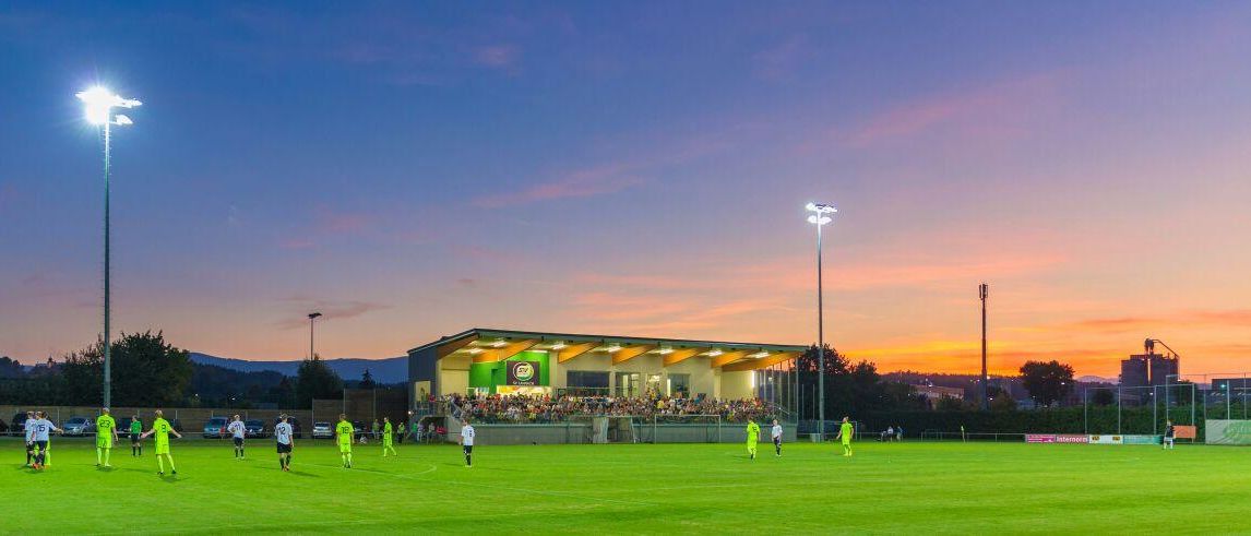 Ein Fußballspiel bei Nacht mit Spielern auf dem Feld, beleuchtet von Flutlichtern, und einer Menge in den Rängen.
