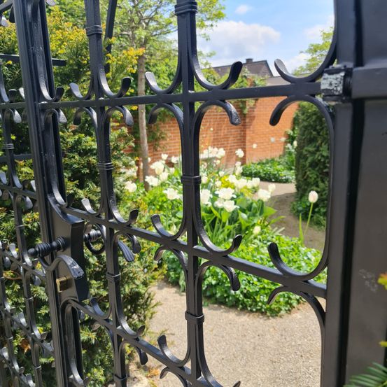 Schwarzes Metalltor mit kunstvollen Verzierungen, das zu einem Garten mit weißen Tulpen und grünen Pflanzen an einem sonnigen Tag führt.
