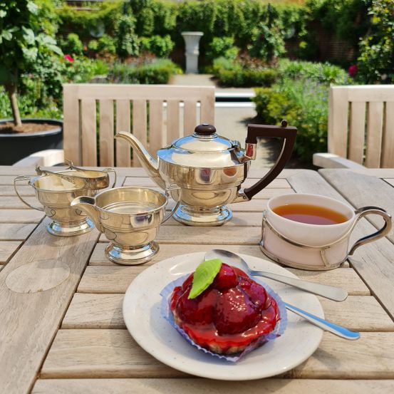 Ein Holztisch mit einem Teeservice, bestehend aus einem Teekessel, einer Tasse Tee und einem Dessert auf einem Teller mit einem Löffel, vor dem Hintergrund eines Gartens.
