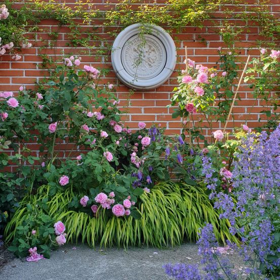 Ein Garten mit einer Ziegelmauer, die mit rosa Rosen, lila Blumen und grünen Blättern geschmückt ist.