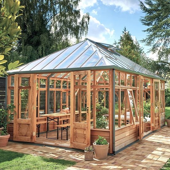 Ein holzverkleidetes Gewächshaus mit Glasdach, umgeben von Topfpflanzen und Bäumen an einem sonnigen Tag.