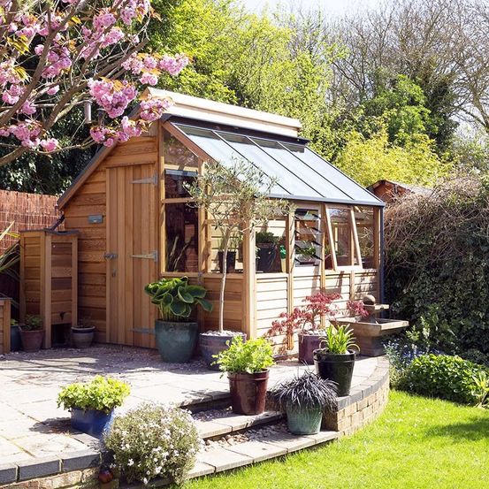 Ein hölzerne Gewächshaus mit Glasdach und Glasfenstern ist von einem Garten mit mehreren Topfpflanzen und Bäumen umgeben.