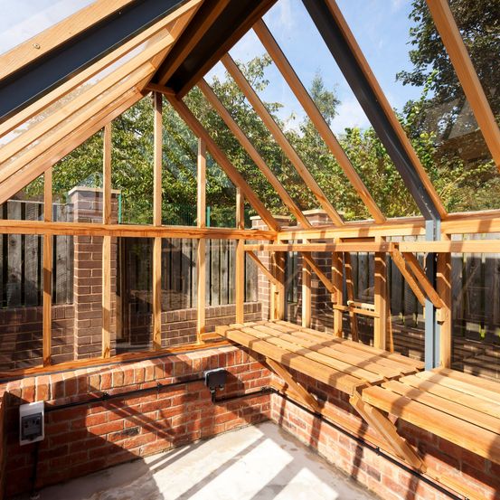 Der Innenraum eines Gewächshauses mit Holzrahmen, Glasfenstern und einer Ziegelwand ist zu sehen.