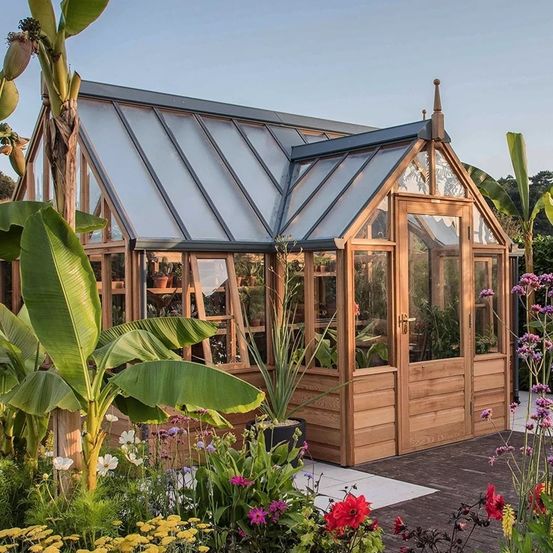 Ein Gewächshaus umgeben von Pflanzen, Blumen und Bäumen mit einem klaren Dach und einer hölzernen Tür und Fenstern.