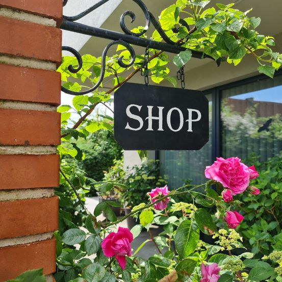 Ein Schild mit dem Wort Shop hängt an der Seite einer Ziegelmauer, umgeben von grünen Pflanzen und rosa Rosen.