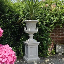 Eine große weiße Keramikurne mit einem dekorativen Henkel steht auf einem Steinsockel in einem Garten, umgeben von grünem Laub und rosa Blumen.