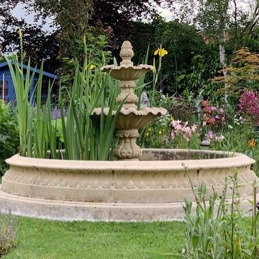 Ein großer Steinbrunnen mit dreistufigem Design befindet sich in der Mitte eines üppigen Gartens, umgeben von verschiedenen Blumen und Pflanzen.
