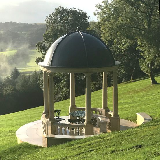 Ein Pavillon mit einer Kuppel steht auf einem Rasenhügel, umgeben von Bäumen.