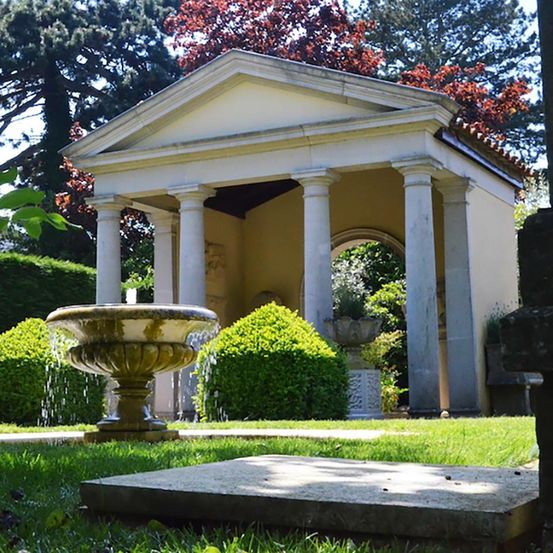 Ein Wasserbrunnen vor einem kleinen Gebäude mit vier Säulen und einem Bogeneingang. Das Gebäude ist von grünen Sträuchern und Bäumen umgeben.