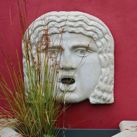 Eine Betonwandskulptur, die ein Gesicht mit einem Ausguss darstellt, umgeben von einer grünen Pflanze, an einer roten Wand.