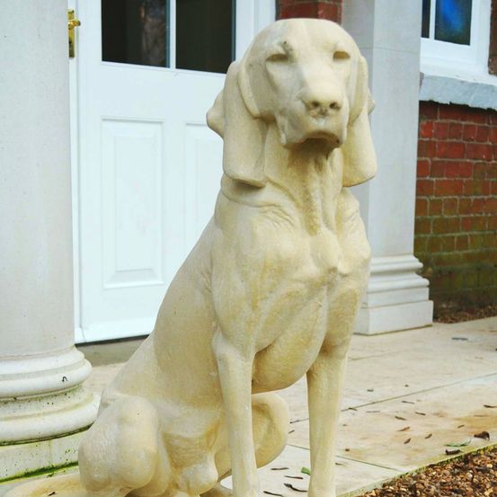 Eine weiße Steinstatue eines sitzenden Hundes steht auf dem Bürgersteig vor einem weißen Gebäude mit einer Ziegelwand.