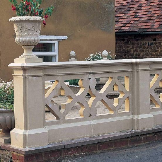 Eine Nahaufnahme einer dekorativen Betonbrüstung mit komplizierten Mustern, die auf einem Ziegelstein-Gehweg ruht. Hinter der Brüstung befindet sich ein Haus mit einem braunen Dach und einem Fenster.