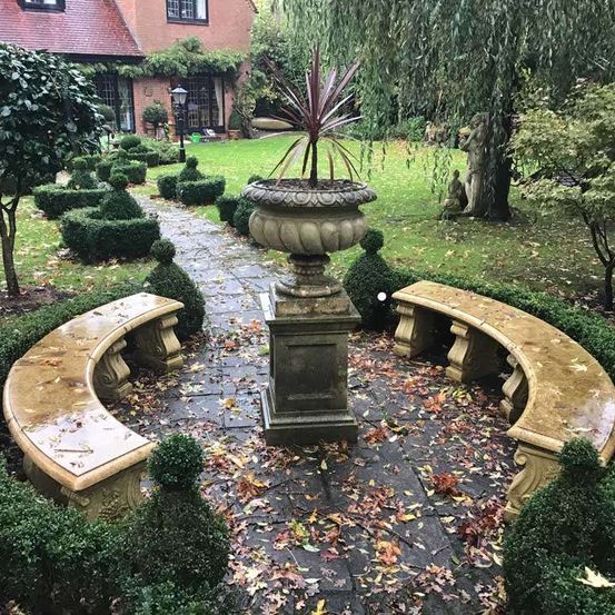 Ein Gartenweg führt zwischen zwei gebogenen Steinbänken hindurch. In der Mitte des Weges steht eine große Steinurne mit einer Pflanze. Der Garten ist von Hecken und Grünpflanzen umgeben, mit einem Haus im Hintergrund.