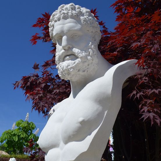 Eine weiße Statue eines bärtigen Mannes mit einem roten Busch im Hintergrund