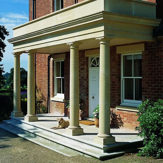 Ein Haus mit einem Vorbau und Säulen, einer weißen Tür und Fenstern, einem Hund, der auf der Veranda liegt, und einigen Pflanzen davor.