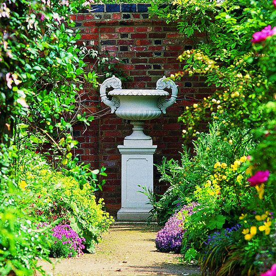 Eine große weiße Steinurne steht inmitten eines üppigen Gartens mit verschiedenen bunten Blumen.