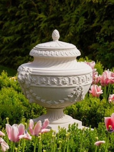 Eine große, weiße Urne mit kunstvollen Schnitzereien steht in einem Garten, umgeben von rosa Blumen und grünen Büschen.