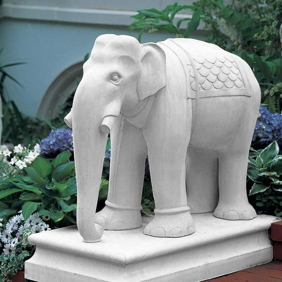 Eine weiße Elefantenstatue mit Sattel steht auf einem Podest inmitten eines Gartens mit blauen und violetten Blumen.