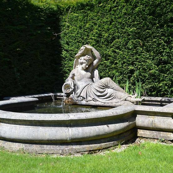Eine Steinskulptur einer liegenden Frau, wahrscheinlich eine Nymphe, in einem Gartenbrunnen.