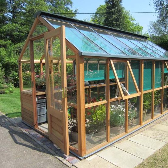 Ein Gewächshaus mit Glasdach und Holzrahmen steht in einem Garten mit grünen Bäumen und Rasen.