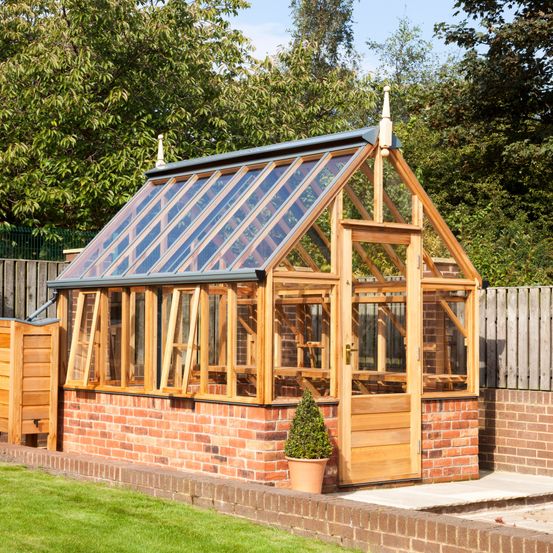 Ein Gartenhaus aus Holz mit Glasfenstern und einer Holztür steht auf einem Ziegelfundament, mit einem Topfpflanzen vor und umgeben von einem Holzzaun.