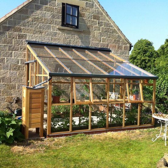 Ein Holzgewächshaus mit Glasdach und -wänden, an ein Steingebäude angebaut, umgeben von Pflanzen und Bäumen, mit einem kleinen Holzschrank auf der linken Seite.
