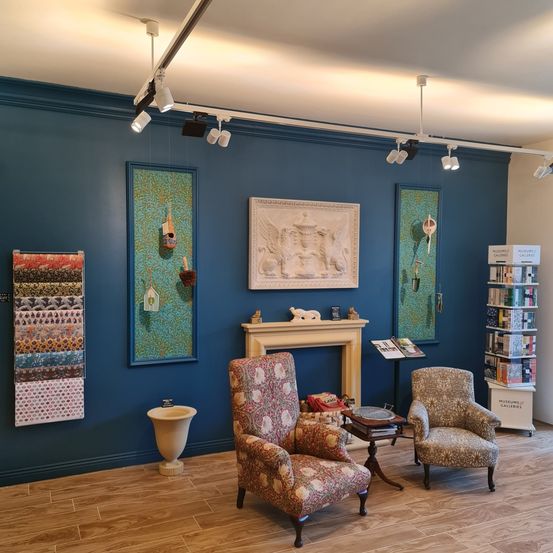 Ein gemütliches Zimmer mit blauen Wänden und Holzböden. Es gibt zwei Sessel mit Blumenmustern, einen Kamin und ein Regal mit Büchern. Über dem Kamin befindet sich ein großes Gemälde. Auf der linken Seite ist eine Wand mit verschiedenen dekorativen Objekten.