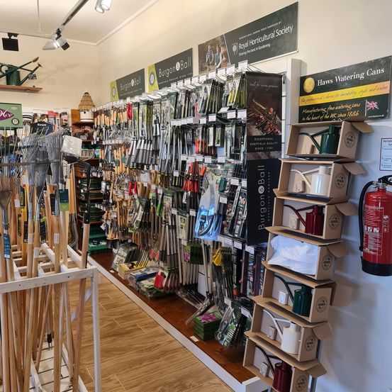 Im Inneren eines Gartenwerkzeugladens mit Holzböden und -wänden. Verschiedene Werkzeuge, darunter Rechen, Schaufeln und Gießkannen, sind an Gestellen und Regalen ausgestellt. Ein roter Feuerlöscher ist an der Wand montiert.