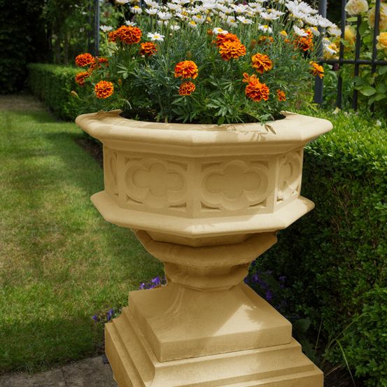 Ein beigefarbener Pflanzkübel mit kunstvollen Schnitzereien auf einer Terrasse, gefüllt mit orangen und weißen Blumen vor dem Hintergrund von Hecken und einem Rasen.