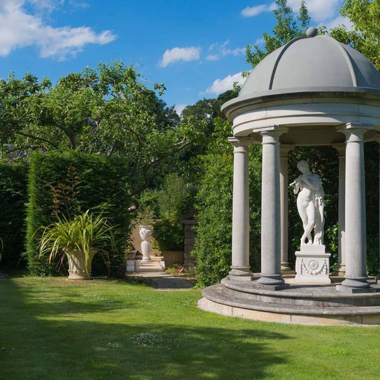 Ein Bild einer Statue einer Frau mit einer Flöte unter einem Pavillon mit Kuppeldach in einem Garten mit grünem Gras und Büschen.